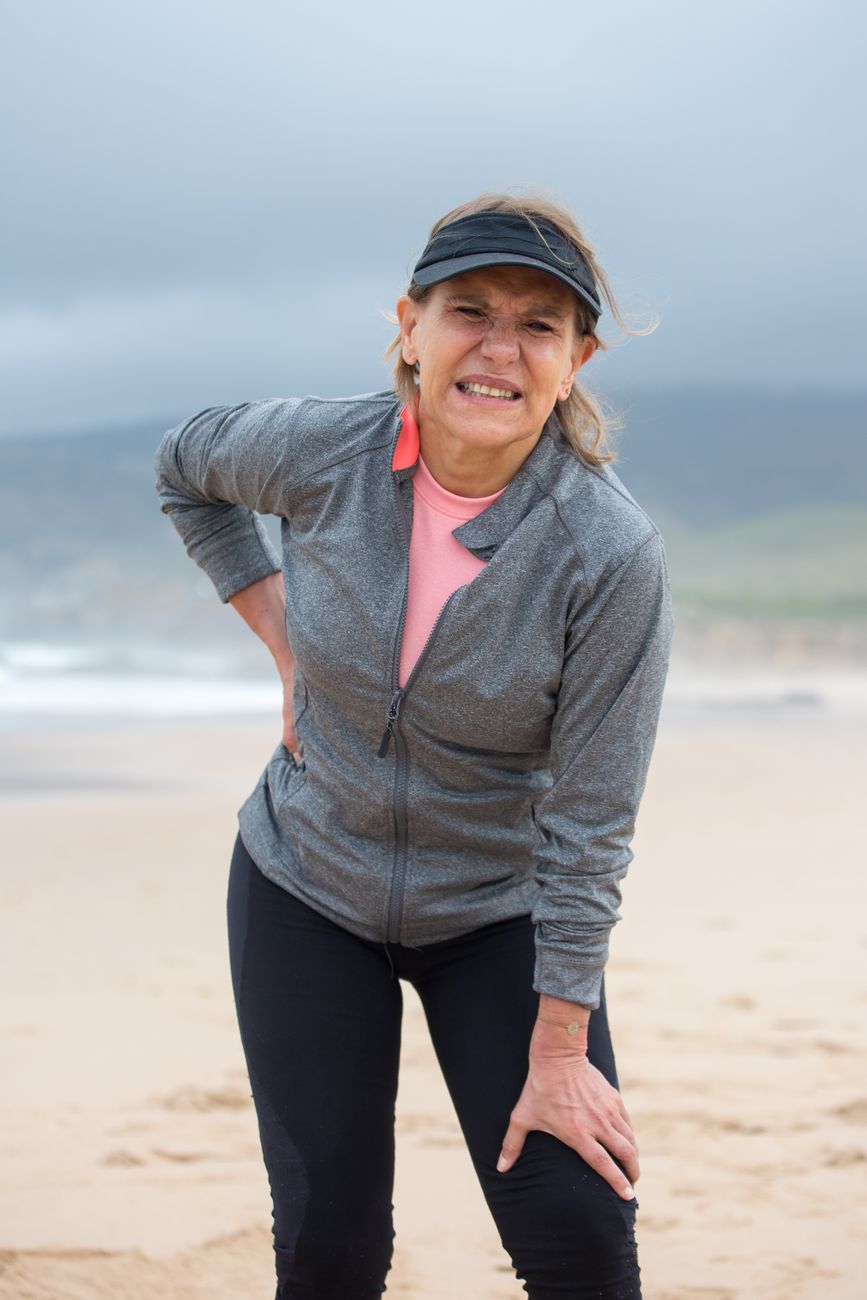 An older woman outdoors on a beach path, pausing with her hands on her hips